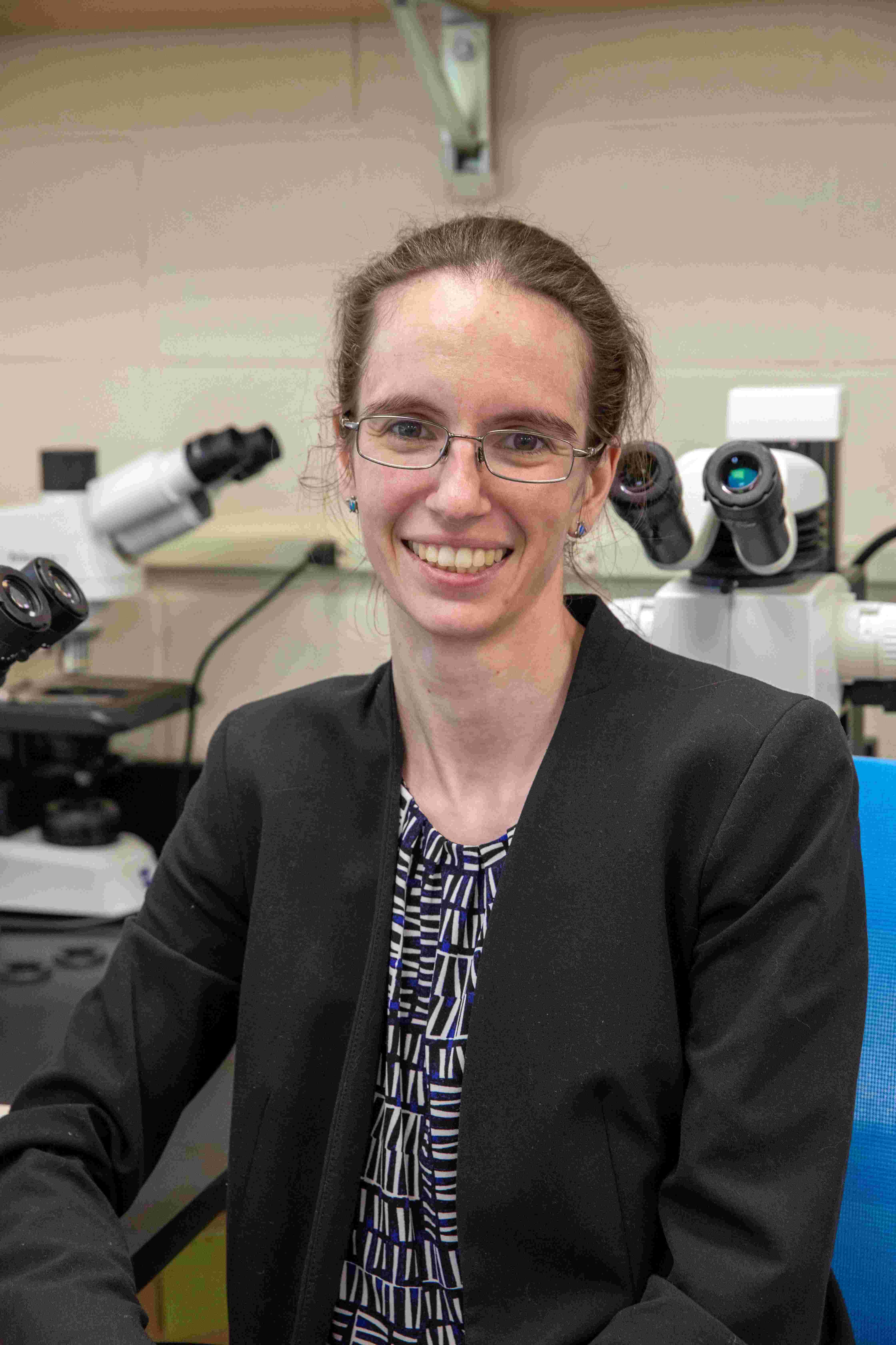 Elizabeth Sibert, a small white woman with brown hair and glasses, is seated in her lab next to several microscopes. She is wearing a blazer and smiling at the camera for this lab-based headshot photo